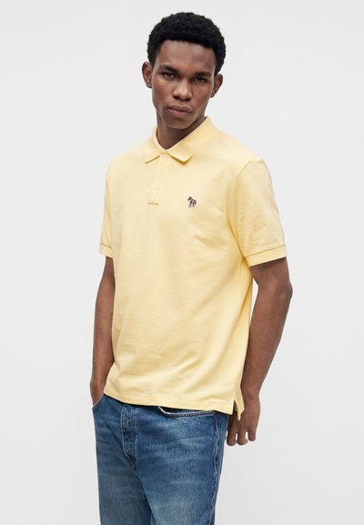 Homme portant un polo jaune pâle avec un petit logo zèbre et un jean bleu, debout les mains dans les poches devant un fond uni.