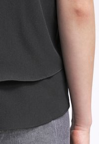 Gros plan sur le bras d’une personne à côté d’un haut en tissu noir superposé et d’un pantalon gris, montrant la texture détaillée et les coutures.