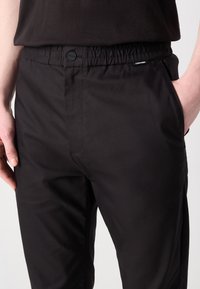 Svarte casual bukser med elastisk midje, knappelukking, sidelommer og en glatt tekstur. Har en liten logo på midjebåndet.