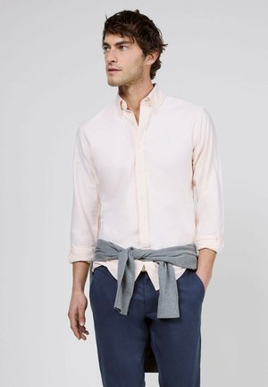 Joven con una camisa rosa clara abotonada con las mangas remangadas, pantalones azul marino y un suéter gris atado alrededor de la cintura, mirando hacia la izquierda.