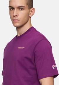 Lilla bomulds t-shirt med rund halsudskæring, der har et lille broderet logo i guld på forsiden og et logo-patch på højre ærme.