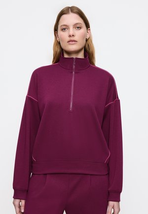 Bordeaux zip-up sweatshirt med høj krave, afslappet pasform og lyserøde sting langs skuldrene og siderne. Blød stofstruktur.