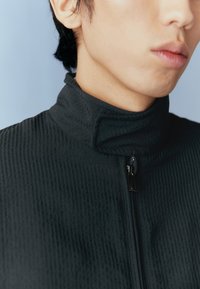 Fekete texturált dzseki magas gallérral és cipzáros elejével. Bordás mintázattal és sima anyagból készült. Közeli kép a nyakkivágás területéről.