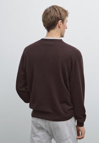 Maglione marrone a maglia con scollo rotondo e spalle scese, visto da dietro, abbinato a jeans grigio chiaro.