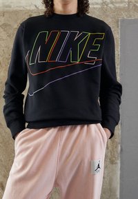 Svart sweatshirt med flerfärgad "NIKE"-logotyp och vita detaljer, bärs med ljusrosa sweatpants med en Jordan-logopatch.