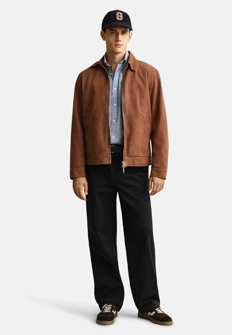 Giacca in suede beige con chiusura a zip, camicia button-up azzurro chiaro, pantaloni neri e sneaker marroni con accenti crema. Cappellino blu navy con logo.