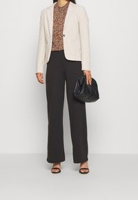Beige Blazer über einer braun gemusterten Bluse, kombiniert mit schwarzen weiten Hosen und einer schwarzen strukturierten Handtasche. Schwarze Öppen-Toe-Schuhe.