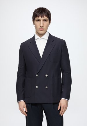 Jeune homme portant un blazer sombre croisé avec des boutons argentés par-dessus une chemise blanche sur un fond clair uni.
