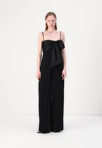 Combinaison noire avec un corsage ajusté et un grand n�œud décoratif à l'avant. Présente des fines bretelles et un pantalon à pattes larges. Texture de tissu lisse.