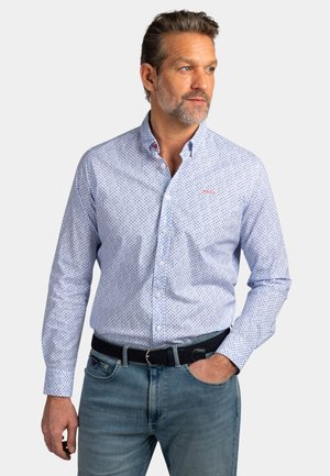 Man met grijs haar en baard, gekleed in een lichtblauw patroonoverhemd met knopen, ingestopt in een blauwe spijkerbroek met een zwarte riem, staand met één hand in zijn zak.