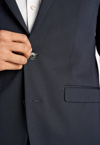 Granatowa marynarka ze strukturalnej tkaniny; posiada zapięcie na dwa guziki, boczną kieszeń oraz kontrastowe przeszycia na klapie.