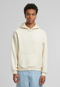Cremefarvet hættetrøje med en frontlomme, snørrelukning i huen og hvid tekst "WHAT GOES AROUND." Blødt stof med en afslappet pasform. Lyst blå jeans.