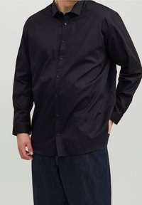 Maglietta nera a maniche lunghe realizzata in cotone morbido, con colletto a polo e tasca laterale, indossata su pantaloni in denim scuri.