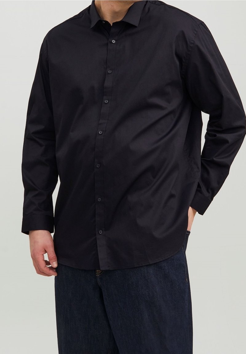 Maglietta nera a maniche lunghe realizzata in cotone morbido, con colletto a polo e tasca laterale, indossata su pantaloni in denim scuri.