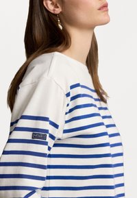 Young woman wearing a white and blue striped long-sleeve shirt with a Polo RL&Co patch on the sleeve and a dangling earring.