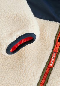 Chaqueta de forro polar con textura cremosa, ribete azul marino y forro rojo. Presenta una cremallera naranja con marca y un tejido verde contrastante.