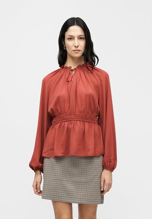 Rød, langærmet bluse med en indsnøret halsudskæring og talje, lavet af glat stof. Parret med et ternet skørt.