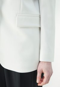 Blazer bianco con un design aderente, texture liscia e una tasca a toppa sul petto. La manica ha un aspetto sartoriale, abbinata a un tessuto nero sottostante.