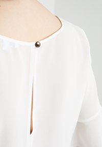 Weiße Bluse mit rundem Halsausschnitt, Schlitz hinten und kleiner metallener Knopfverschluss im Nacken. Glattes, leichtes Material mit weicher Textur.