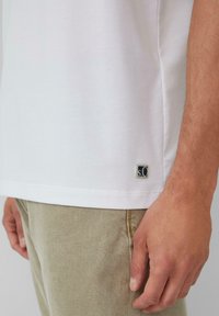 Weißes Baumwoll-T-Shirt mit kurzen Ärmeln, mit einem kleinen blauen und grauen Logo-Tag in der unteren linken Ecke. Beigefarbene Hose mit strukturiertem Finish.