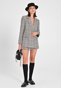 Blazer a doppio petto grigio a quadri abbinato a una mini gonna coordinata, calze nere sopra il ginocchio e una borsa nera con texture e dettagli dorati.