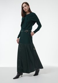 Frau in einem dunkelgrünen Langarmoberteil und passendem Rock mit unterer Lederpartie und schwarzem Gürtel, kombiniert mit schwarzen Knöchelstiefeln.