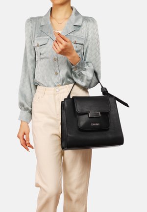Borsa a tracolla in pelle nera con una forma strutturata, caratterizzata da una chiusura a patta frontale e un manico con dettagli in evidenza. Indossata da una persona con una camicia chiara.