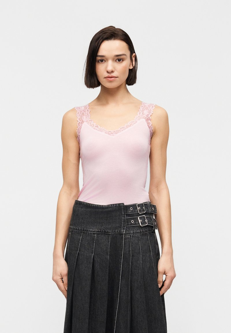 Νεαρή γυναίκα με κοντά σκούρα μαλλιά που φοράει ροζ τοπ με δαντέλα και μαύρη πλισέ φούστα με διπλές αγκράφες.