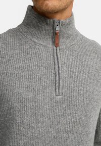 Grå ribbet sweater med høj krave og lynlås foran. Har en brun læder trækklap og en struktureret stofoverflade hele vejen rundt.