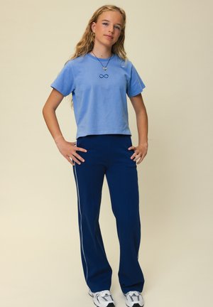 Tienermeisje met lang blond haar staat met de handen in de zij, draagt een lichtblauw t-shirt, donkerblauwe broek met witte strepen en witte sneakers.