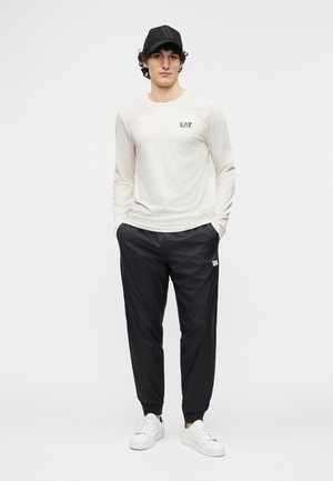 Άνδρας που φοράει λευκό μακρυμάνικο πουκάμισο με το λογότυπο «EA7», μαύρο υφασμάτινο παντελόνι jogger, λευκά αθλητικά παπούτσια και μαύρο καπέλο με ανάγλυφο λογότυπο «AX».