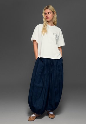 Blondína stojí s rukami vreckách, má na sebe voľné biele tričko s textom a široké tmavomodré nohavice, hnedé topánky, na šedom pozadí.