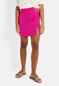 Minifalda fucsia con una abertura frontal y detalles en dorado. Combinada con sandalias deslizantes de estampado de leopardo. Textura suave y diseño ajustado.
