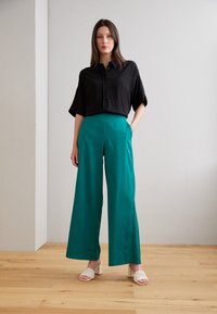 Svart skjorte med korte ermer, sammen med høyt liv turkise vide bukser. Hvite blokkhevede sandaler med en teksturert design.