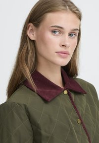Quiltet olivengrøn jakke med en burgundy kordfløjlskrave. Har guldknaplukning og et diamantmønster på stoffet.