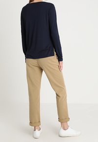 Personne debout de dos portant une chemise bleu marine à manches longues, un pantalon beige retroussé et des baskets blanches sur un fond uni.