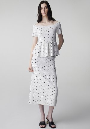 Frau trägt ein weißes schulterfreies Peplum-Oberteil und einen passenden Rock mit Polka-Dots, kombiniert mit schwarzen offenen Sandaletten mit Absatz, steht vor einem grauen Hintergrund.