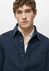 Navy blue jacket with a pointed collar, front buttons, and two chest pockets, worn over a white collared shirt, featuring a smooth texture.