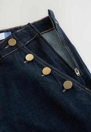 Pantalones vaqueros de mezclilla azul oscuro con cremallera, tres grandes botones de latón, costuras en contraste y una textura suave.