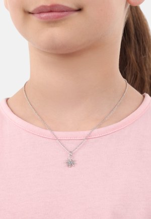 Silberne Kettenhalskette mit einem Schneeflockenanhänger, der türkisfarbene Akzente hat, getragen über einem hellrosa T-Shirt mit rundem Ausschnitt.