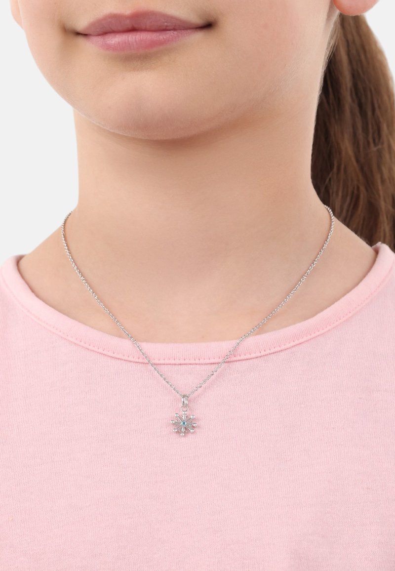 Silverfärgad kedjehalsband med en snöflingeberlock som har turkosa detaljer, bärs över en ljusrosa t-shirt med rund halsringning.