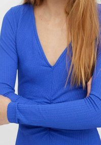 Blaues Langarmoberteil mit V-Ausschnitt, strukturiertem Stoff und gerafftem Detail an der Taille, mit eng anliegenden Ärmeln und glatter Oberfläche.