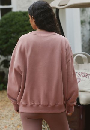 Femeie cu păr lung și închis la culoare, purtând un hanorac roz prăfuit supradimensionat și colanți asortați, stând afară aproape de o grădină și un scaun deschis la culoare.