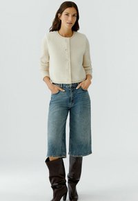 Crème-kleurige boucle cardigan met ronde hals, parelknopen en lange mouwen, gecombineerd met lichtblauwe wijde jeans en bruine kniehoge laarzen.