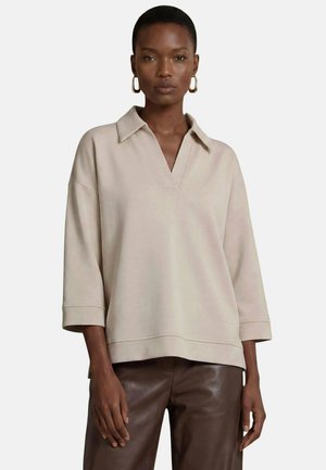 Kvinde iført beige langærmet skjorte med krave og brune læderbukser, med øreringe i form af ringe, stående mod en ensfarvet lys baggrund.