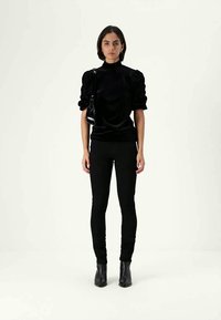 Γυναίκα στέκεται με το πρόσωπο μπροστά, φορώντας μαύρη μπλούζα με φουσκωτά μανίκια, μαύρο παντελόνι, μαύρες μποτάκια μέχρι τον αστράγαλο και μια μικρή μαύρη τσάντα ώμου.