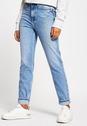 Person, die hellblaue Jeans mit geradem Bein und Umschlägen sowie weiße Sneaker trägt und vor einem schlichten weißen Hintergrund steht.