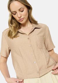 Short-sleeve, collared shirt in light beige fabric. Features a front pocket and five buttons down the front with a textured finish.