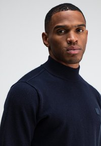 Marineblauer Rollkragenpullover aus strukturiertem Stoff, mit einem gerippten Kragen und einem kleinen Logo-Patch auf der linken Brust.