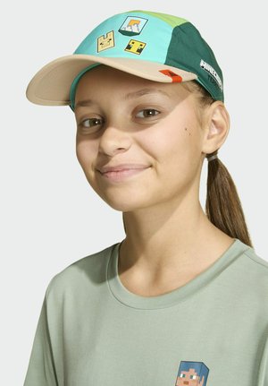 Persona joven con una gorra verde temática de Minecraft y una camiseta de un verde claro a juego, sonriendo levemente con el cabello recogido.